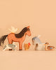 The Ostheimer Handmade Wooden Farm Animals Set, featuring seven handcrafted wooden animals made from sustainably sourced materials—a horse, a cat, a goat, a chicken, two ducks, and a bird perched on the horse—is displayed on a plain light brown background.