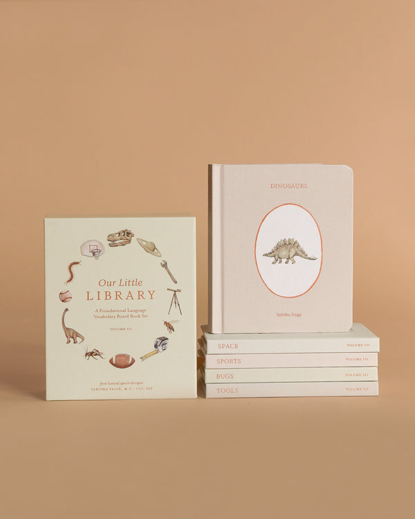Two stacks of books on a beige surface: one upright, "Our Little Library Vol. 3 (5 Board Books)" with cute animal and object illustrations, and a horizontal stack with striped board books on topics like dinosaurs and space—perfect for baby showers and early language learning.