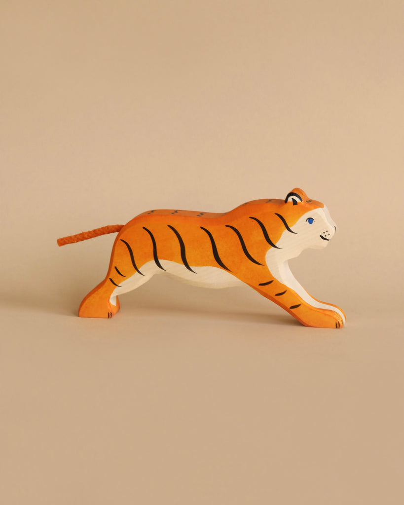 A small, handcrafted wooden tiger figurine with orange and black stripes. The tiger is depicted mid-stride, looking forward with a playful expression. Made in Europe, this Holztiger Tiger, Running figure is set against a plain beige background, highlighting its beautiful craftsmanship.