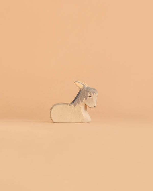 The Ostheimer Donkey - Resting, a small and minimalist wooden figurine with gray ears and mane, reminiscent of classic handcrafted toys, sits on a smooth, light peach background.