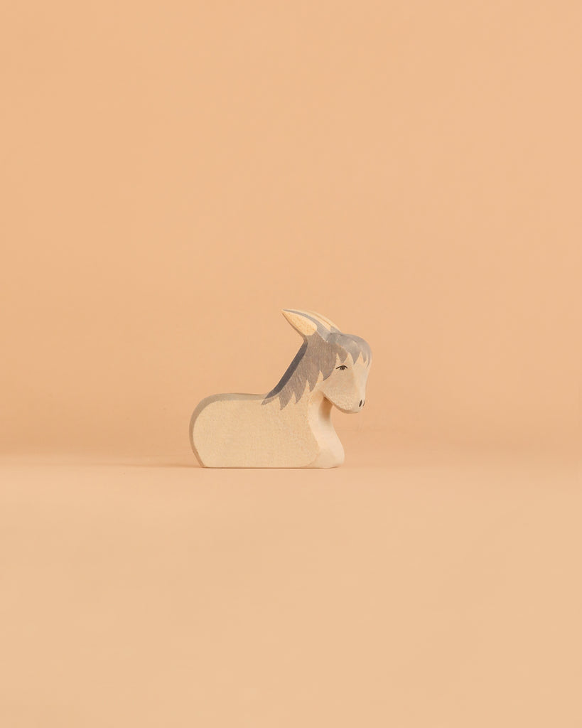 The Ostheimer Donkey - Resting, a small and minimalist wooden figurine with gray ears and mane, reminiscent of classic handcrafted toys, sits on a smooth, light peach background.