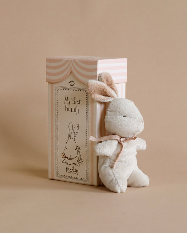 A soft white plush bunny with pink inner ears and a pink ribbon around its neck, standing next to its beige and pink striped giftbox titled "Maileg My First Bunny - Dusty Rose".