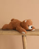 The Steiff Barny Brown Bear Stuffed Animal, a brown plush bear, lies on its stomach across a wooden bench against a plain beige background, evoking the charm of classic Steiff bears.