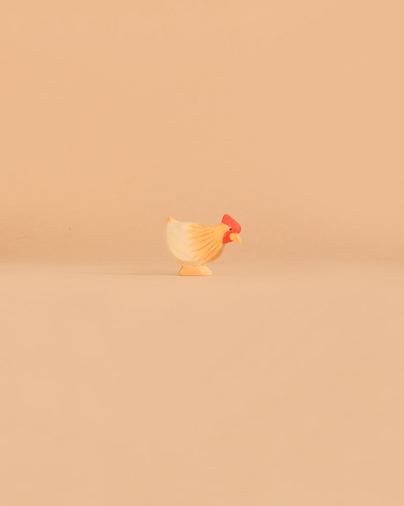 A wooden chicken figurine with a red comb from the Ostheimer Handmade Wooden Farm Animals Set stands on a plain light beige background, reflecting the charming simplicity of these handcrafted toys.