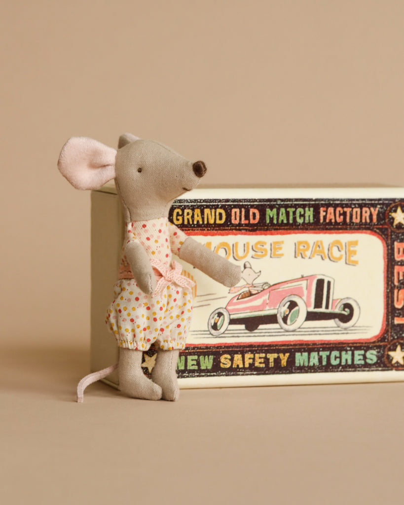 A small, gray Maileg mouse from the Little Sister Bedroom Set stands upright next to a vintage-style matchbox decorated with a red race car and retro text while wearing a floral dress.