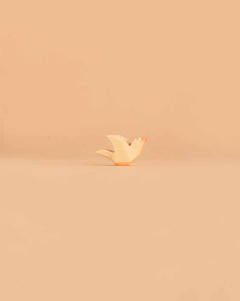 A charming, small, light-colored bird figurine with an orange beak from the Ostheimer Handmade Wooden Farm Animals Set is centered on a smooth, beige surface with a minimalist beige background—an elegant example of handcrafted wooden toys.
