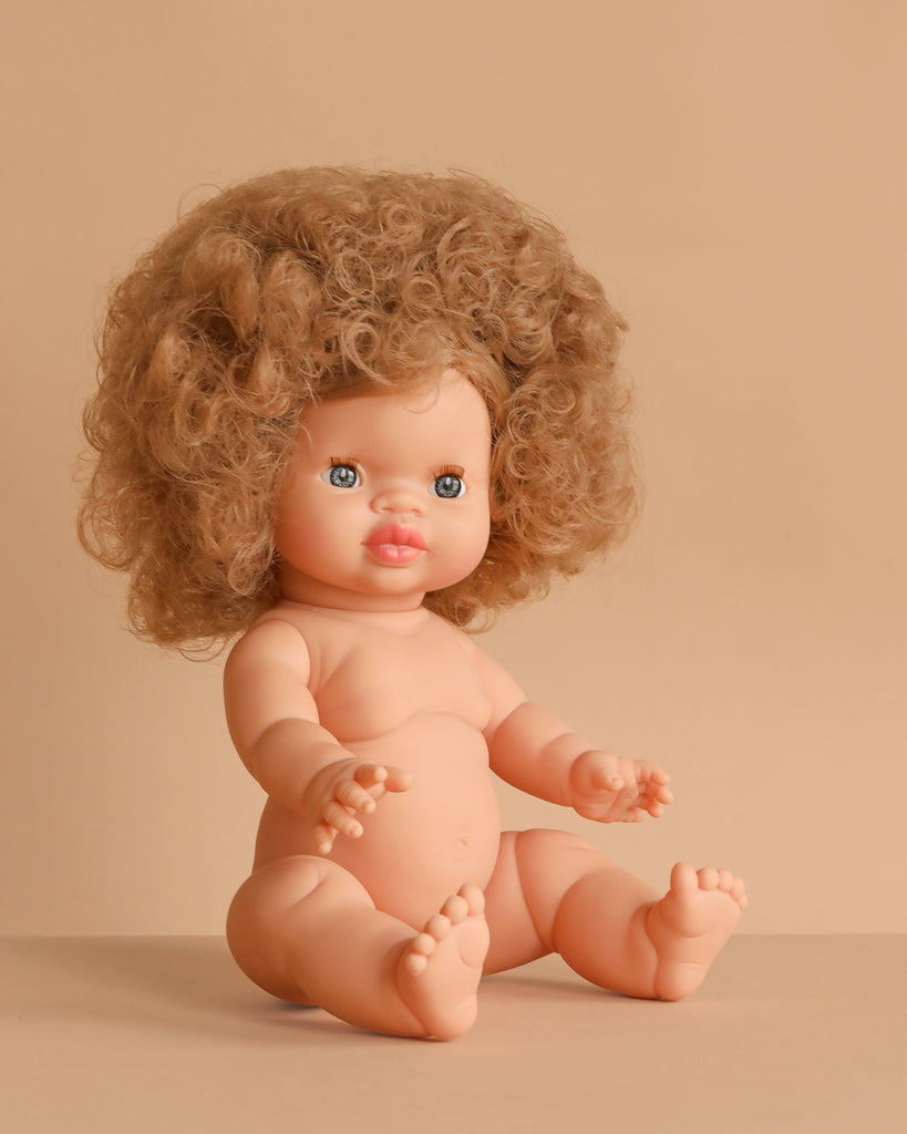 The Minikane Baby Doll (13") – Anais sits on a beige surface with light skin, blue eyes, and a large curly light brown afro. This anatomically correct doll is made in Spain. Clothing sold separately.