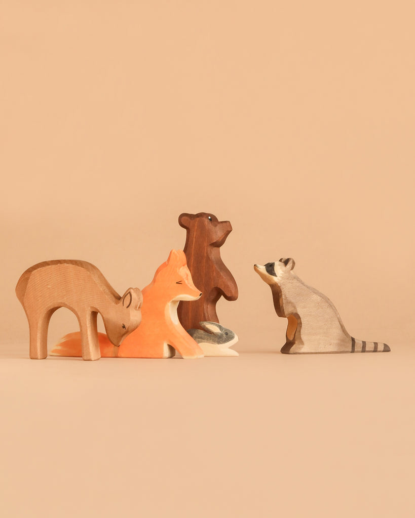 The Ostheimer Handmade Wooden Woodland Animals Set, featuring four handcrafted wooden figurines—a deer, fox, bear, and raccoon—are artfully arranged in a semi-circle on a neutral beige background. Inspired by classic Ostheimer wooden toys, this sustainable set combines timeless design with eco-friendly craftsmanship.