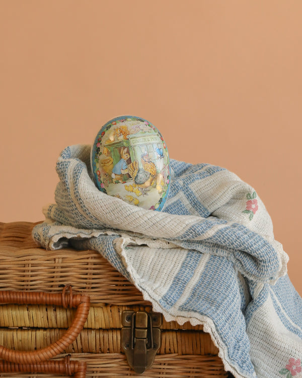A Cardboard Beatrix Potter Egg, Small (Morning at Ginger & Pickles’ Shop) with vintage illustrations rests on a blue and white woven cloth atop a wicker suitcase, set against a peach-colored background.