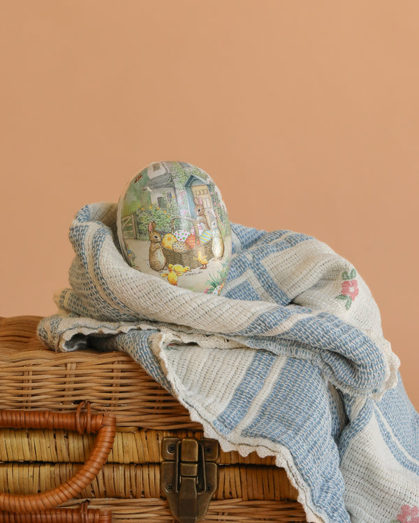 The small Cardboard Beatrix Potter Egg (The Easter Basket Gathering) features rabbits and flowers, resting on a blue-and-white blanket that covers a wicker basket, with a peach background.