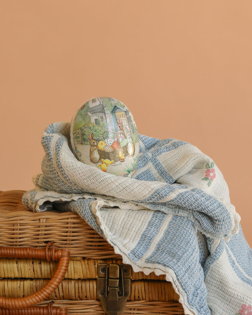 The small Cardboard Beatrix Potter Egg (The Easter Basket Gathering) features rabbits and flowers, resting on a blue-and-white blanket that covers a wicker basket, with a peach background.