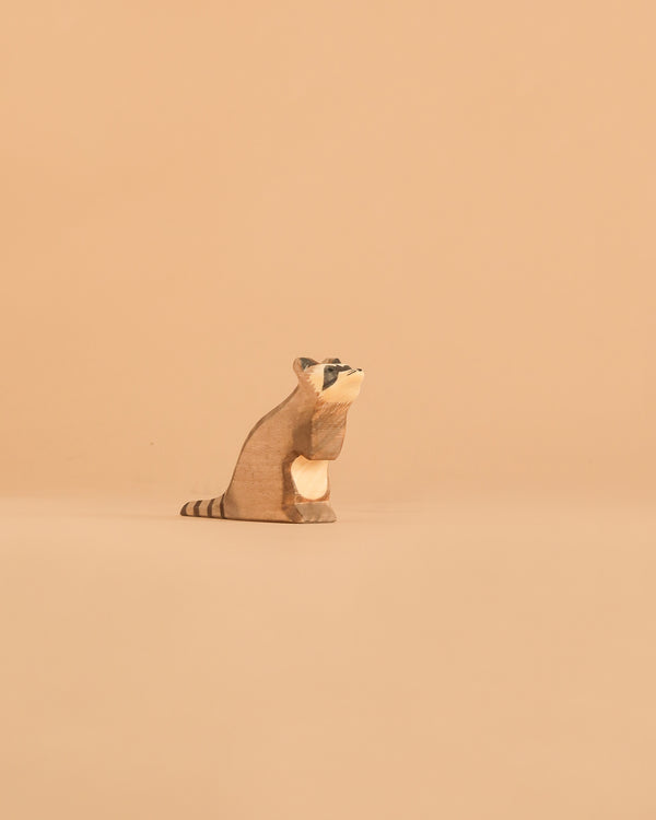 The Ostheimer Raccoon - Sitting, a small handcrafted wooden figurine, rests on a plain beige surface against a matching background, facing slightly to the left. Perfect for imaginative play, this piece reflects the timeless charm of Ostheimer wooden toys.