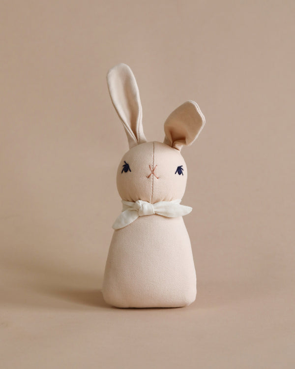 A Polka Dot Club Rabbit Rattle - Peach with long ears and embroidered eyes and nose, wearing an organic cream necktie around its neck, set against a plain beige background.
