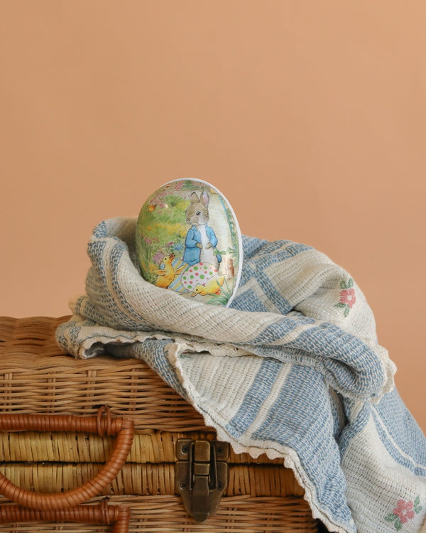 A small Cardboard Beatrix Potter Egg (Rabbit with Ducklings) rests on a blue and white striped cloth, draped over a wicker basket against a plain peach background.