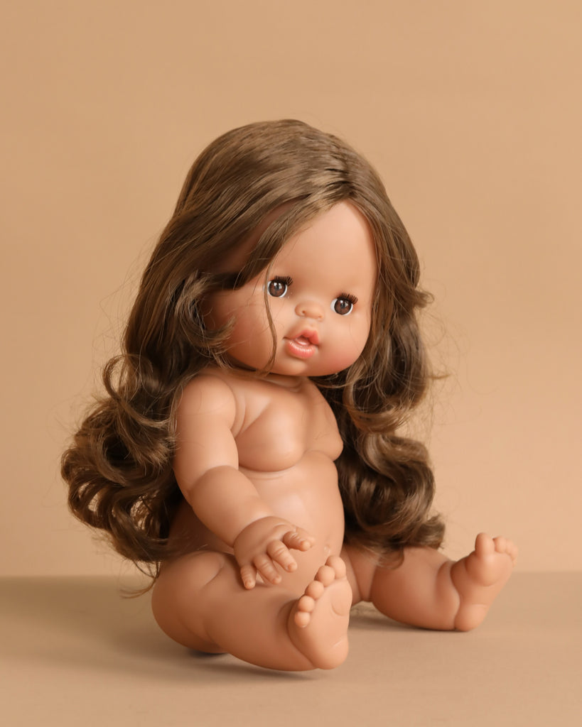 The Minikane Baby Doll (13") - Leopoldine, featuring long wavy brown hair and brown eyes, sits upright on a beige surface against a matching background. Clothing sold separately.