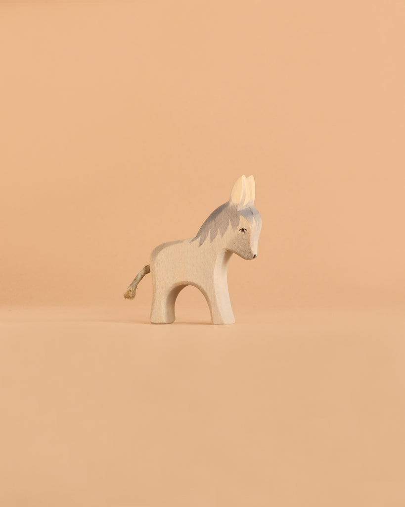 The Ostheimer Donkey - Small is a minimalist wooden figurine inspired by classic Ostheimer designs, featuring painted gray details and a simple rope tail. It stands on a beige surface with a plain beige background—ideal for imaginative play and collectors of handcrafted wooden toys.