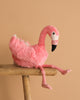 The Steiff Flamingo Stuffed Animal, featuring bright pink plumage and long legs, sits on a wooden stool against a plain beige background.