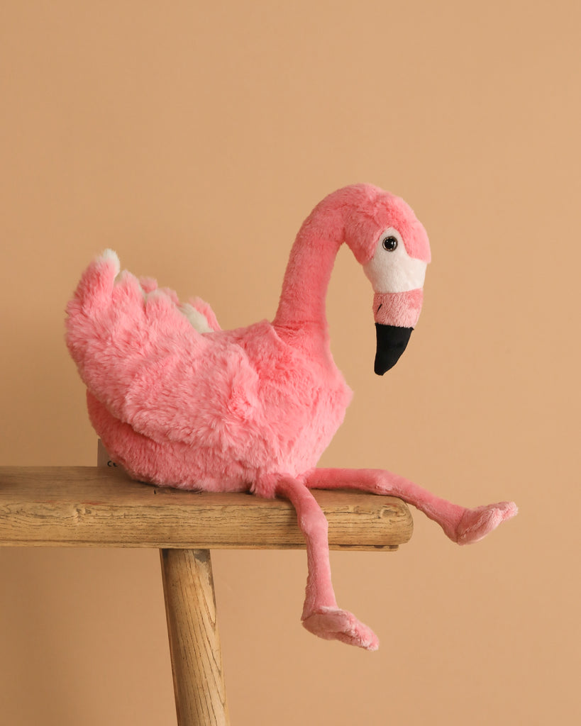 The Steiff Flamingo Stuffed Animal, featuring bright pink plumage and long legs, sits on a wooden stool against a plain beige background.