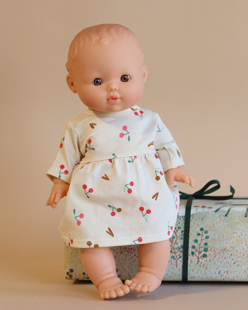 The Minikane Doll (13") - Faustine with Cherry Dress, made from phthalate-free vinyl, is dressed in a white cherry-patterned outfit and is placed on top of a floral gift box wrapped with a green ribbon. The neutral background adds to the gentle, minimalist ambiance.