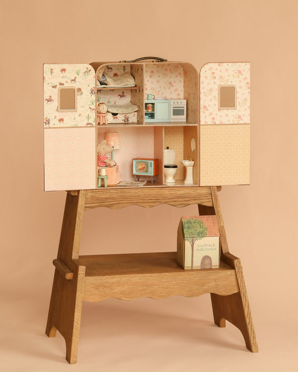The Maileg Suitcasa Furnished Set features a charming wooden dollhouse with two open doors that reveal miniature rooms and furniture, including a kitchen, bathroom, and bedroom decorated with adorable Maileg mouse-sized pieces. Resting on a wooden stand with a book and tiny house beneath, this delightful set invites imaginative play with Maileg dollhouse accessories.