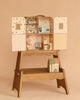 The Maileg Suitcasa Furnished Set features a charming wooden dollhouse with two open doors that reveal miniature rooms and furniture, including a kitchen, bathroom, and bedroom decorated with adorable Maileg mouse-sized pieces. Resting on a wooden stand with a book and tiny house beneath, this delightful set invites imaginative play with Maileg dollhouse accessories.