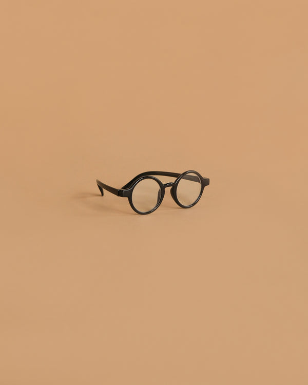A pair of Minikane Doll Accessories Harry Sunglasses in black is centered on a plain, light brown background.