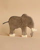 A gray elephant from the Holztiger Handmade Wooden Safari Animals Set, crafted from maple and beech wood, stands on a light beige background. It features white tusks, painted details, and a minimalist design.