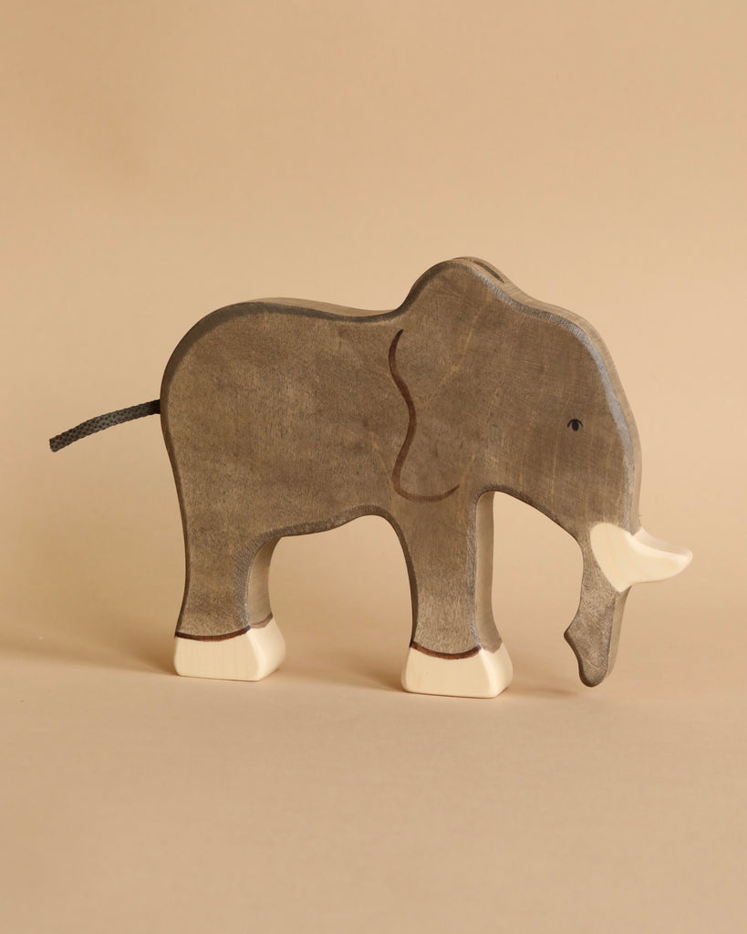 A gray elephant from the Holztiger Handmade Wooden Safari Animals Set, crafted from maple and beech wood, stands on a light beige background. It features white tusks, painted details, and a minimalist design.