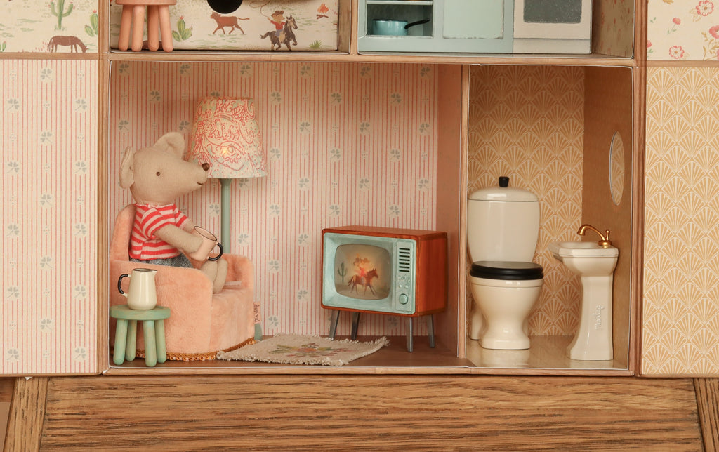 A miniature bear in a striped shirt relaxes in an armchair, watching TV in a dollhouse living room adorned with the charming Maileg Suitcasa Furnished Set. Nearby are a lamp, toilet, sink, side table with a mug, and patterned wallpaper, creating a cozy scene reminiscent of the delightful Maileg mouse set.