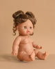 The 13" Minikane Baby Doll With Sleeping Eyes - Madeleine sits on a beige surface with brown hair in two buns. The anatomically correct doll features chubby limbs and a slightly open mouth. Clothing sold separately.