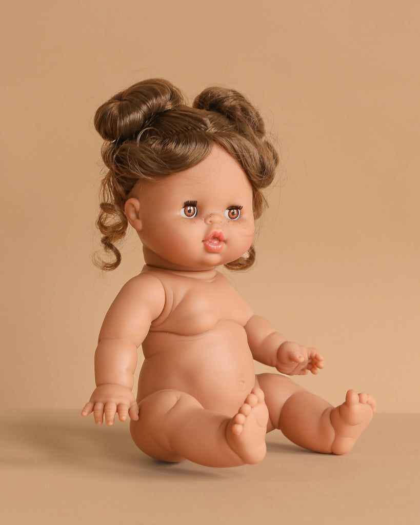 The 13" Minikane Baby Doll With Sleeping Eyes - Madeleine sits on a beige surface with brown hair in two buns. The anatomically correct doll features chubby limbs and a slightly open mouth. Clothing sold separately.
