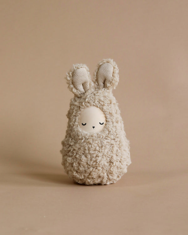 A plush toy resembling a Handmade Musical Roly Poly - Bunny with a beige and cream color palette, standing upright against a plain beige background. The bunny has large, floppy ears and emits soft musical sounds, enhancing its charm.