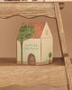 A small house-shaped box featuring a tree illustration and the words "Louimax & Stella Stardust" sits on a wooden shelf, making the perfect addition to any Maileg Suitcasa Furnished Set or serving as charming accessories for your Maileg dollhouse.