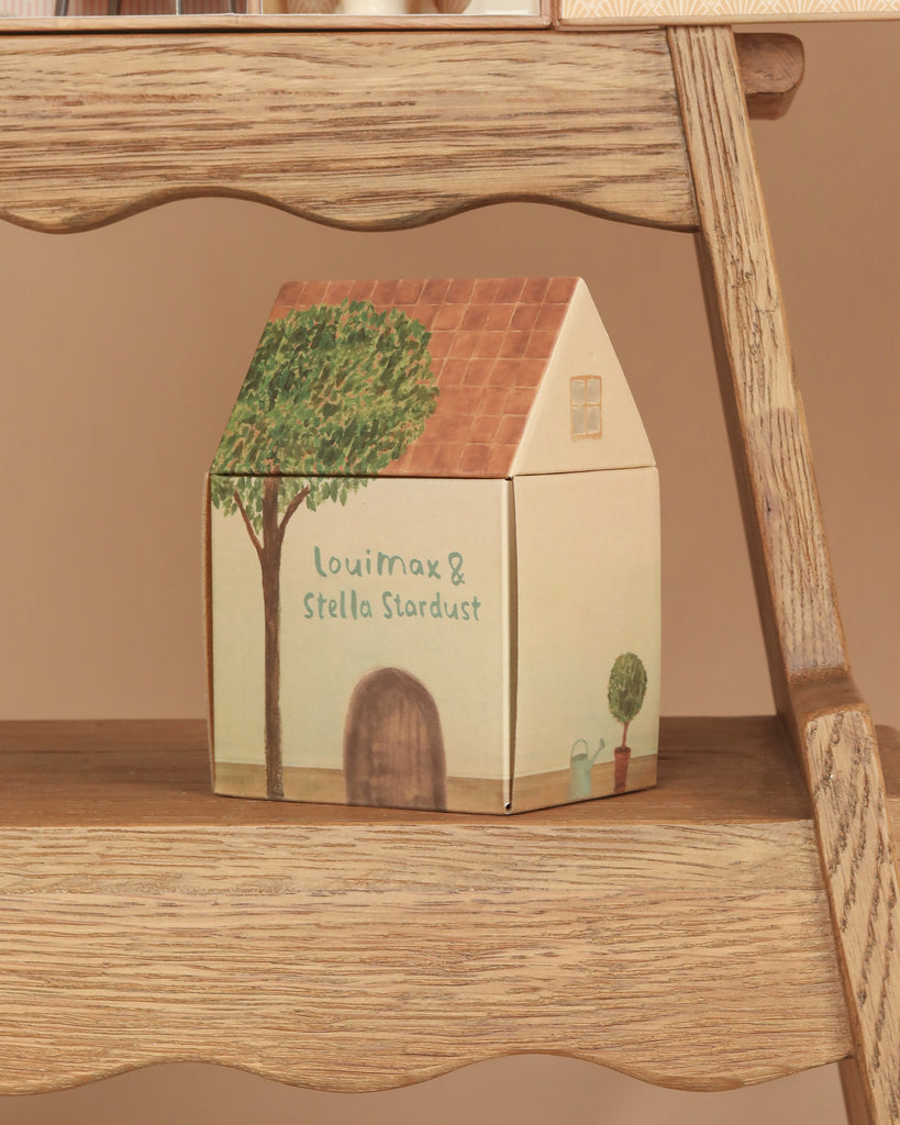 A small house-shaped box featuring a tree illustration and the words "Louimax & Stella Stardust" sits on a wooden shelf, making the perfect addition to any Maileg Suitcasa Furnished Set or serving as charming accessories for your Maileg dollhouse.
