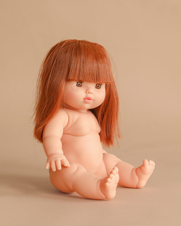 The Minikane Baby Doll (13") - Amber features light skin, long straight red hair with bangs, and sits on a beige surface against a beige background. This adorable doll is made in Spain. Clothing sold separately.