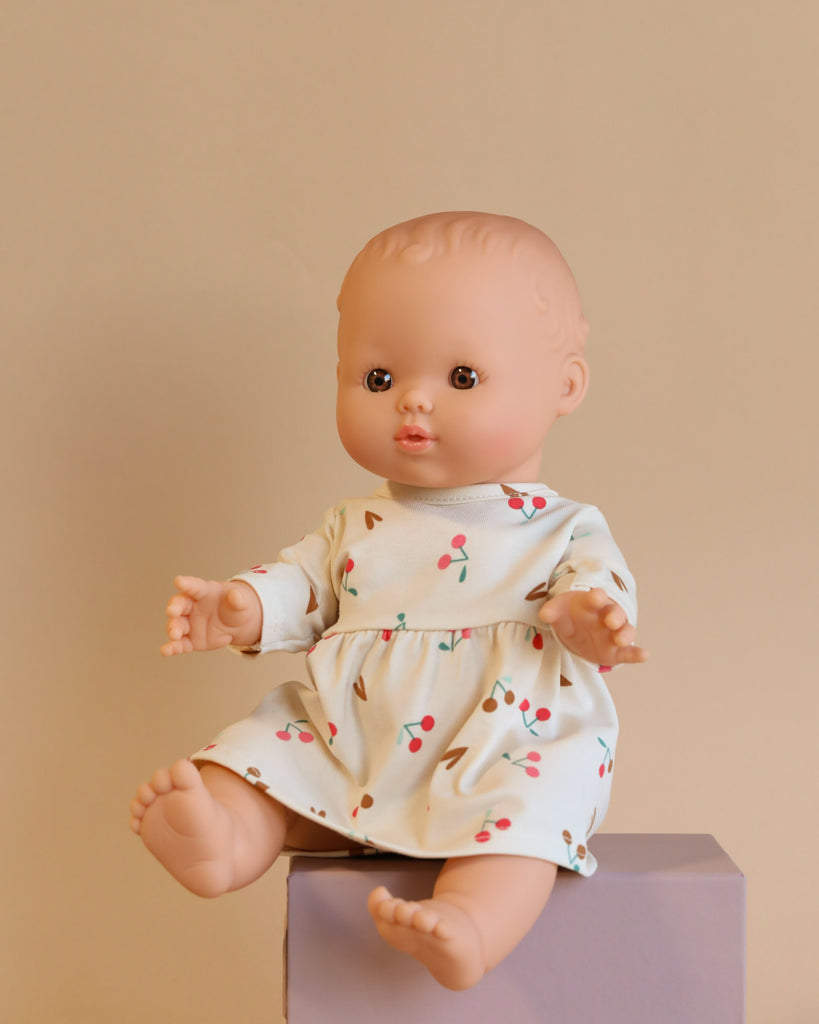 The Minikane Doll (13") - Faustine with Cherry Dress, crafted from phthalate-free vinyl, features light skin and wears a charming white dress with a cherry pattern. Displayed on a light gray box against a beige background, this doll has outstretched arms and a neutral expression, making it ideal for imaginative playtime.