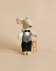 A small Maileg Waiter Mouse is dressed in black trousers, a white shirt, and a black and white checkered vest with a bowtie. The mouse appears to be standing next to a tiny golden chair against a plain beige background.