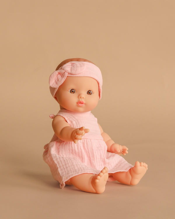 The Minikane Vintage Baby Doll (13") - Faustine with Clothing features light skin, a pink headband with a bow, and a pink dress. Made from phthalate-free vinyl for safe play, Faustine sits on a beige background with one arm raised.