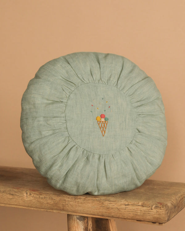 The Maileg Small Round Cushion in Mint features a ruffled edge and embroidered ice cream cone at the center, shown on a rustic wooden stool against a beige background.