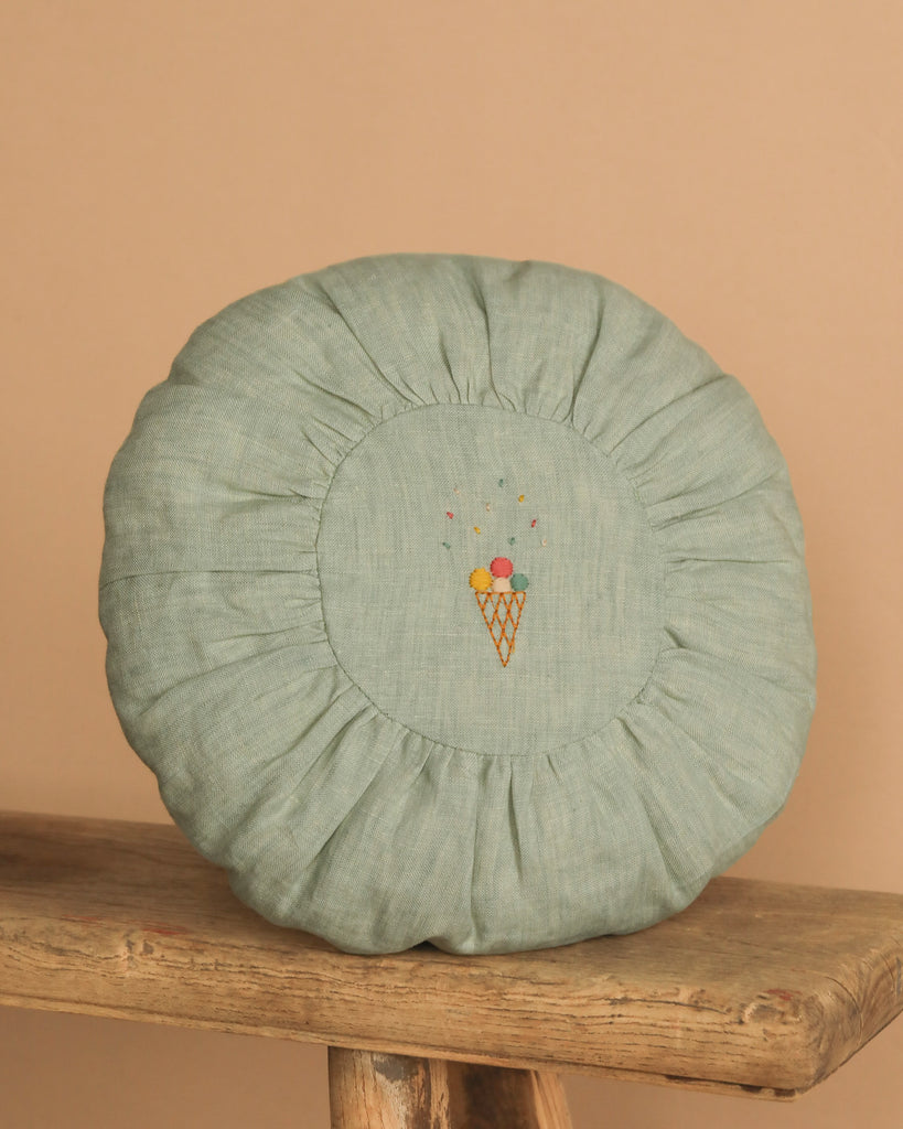 The Maileg Small Round Cushion in Mint features a ruffled edge and embroidered ice cream cone at the center, shown on a rustic wooden stool against a beige background.