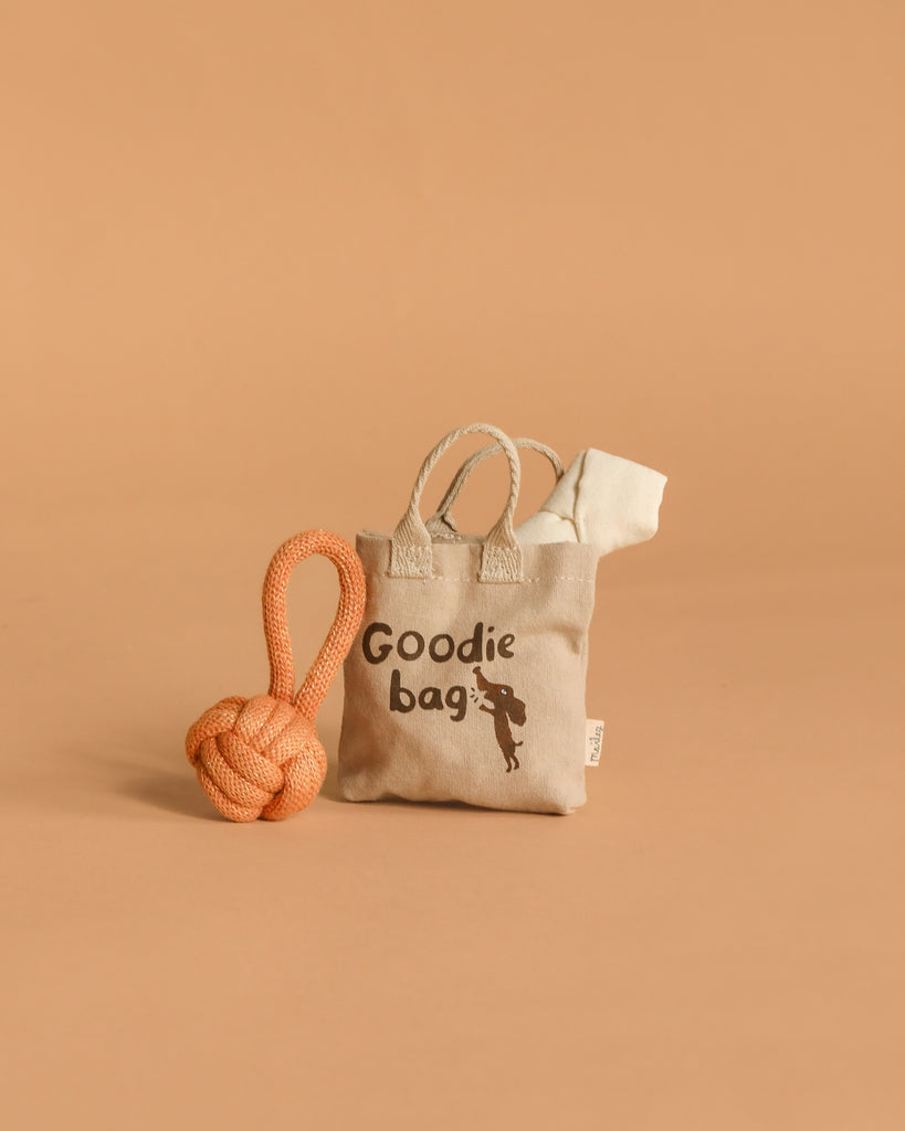 The Maileg Puppy Playtime Set with Goodie Bag includes a beige tote labeled "Goodie bag" with a dog silhouette, an orange rope toy, and a white item—ideal accessories for your Maileg Puppy.