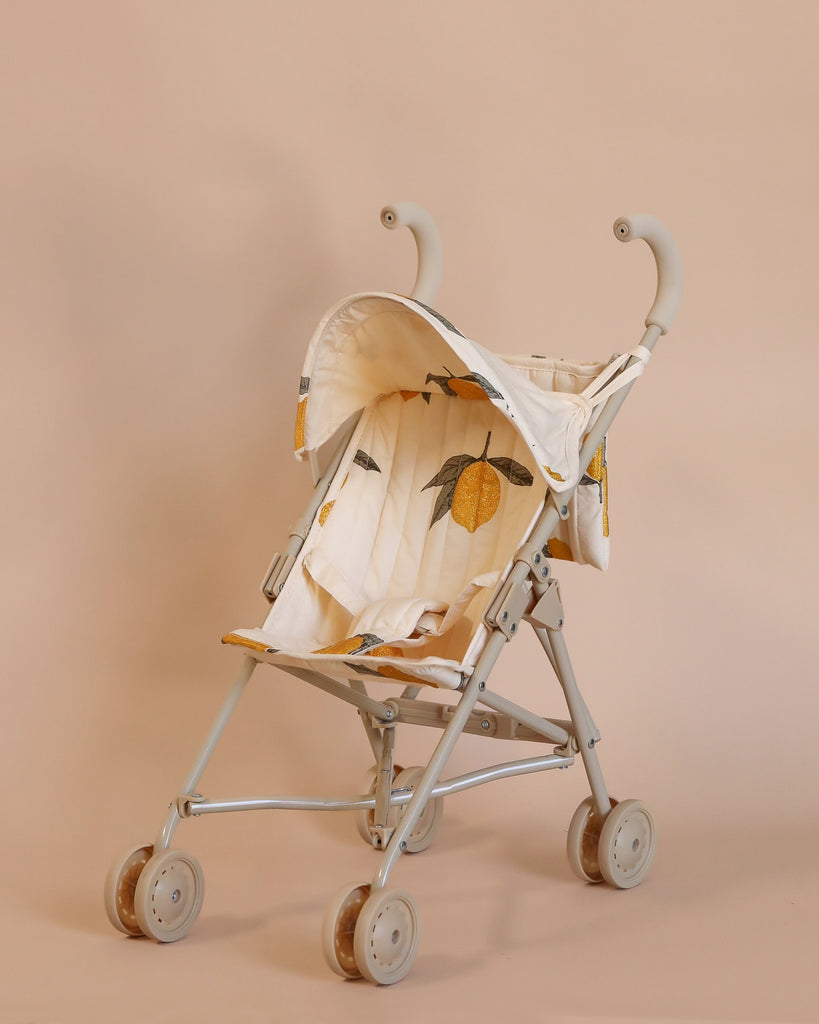 The Konges Sloejd Zoe Doll Stroller - Lemon is a compact and lightweight stroller featuring a beige frame with double wheels. Its fabric seat and canopy are adorned with a pattern of oranges and green leaves, complemented by a foldable shade, against a simple, neutral beige background.