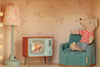 A small toy mouse in a red-striped shirt sits on a blue chair—part of the delightful Maileg Fully Furnished Farmhouse Set + Surprise Gift (ships in one week)—next to a vintage-style lamp and TV in a dollhouse room with floral wallpaper, creating the perfect scene for any Maileg Farmhouse Dollhouse or collection of dollhouse accessories.