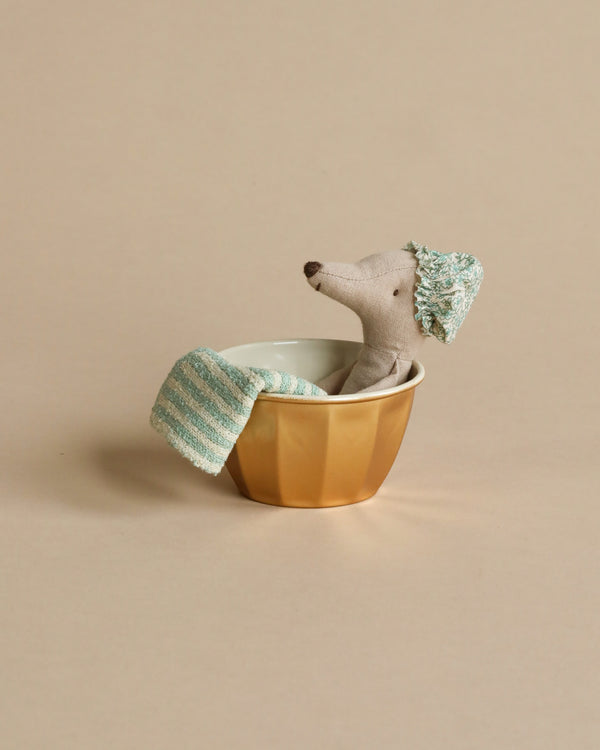 A Maileg Wellness Mouse, Big Sister (Mint) named Ms. Mouse, wearing a striped knit hat and scarf, peeking out from inside a golden bowl against a plain beige background.