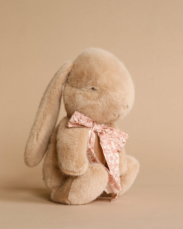 Light brown plush bunny with light pink floral bow around its neck. Beige background. 