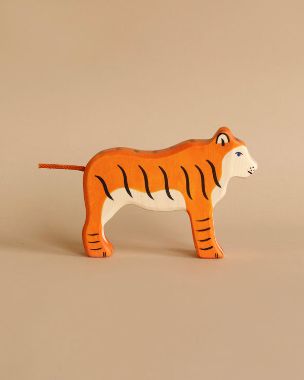 A handcrafted wood toy tiger stands on a beige background. The piece, part of the renowned Holztiger Tiger, Standing figures made in Europe, boasts an orange body with black stripes, a white face and underside, and blue detailing around the eyes.