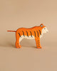 A handcrafted wood toy tiger stands on a beige background. The piece, part of the renowned Holztiger Tiger, Standing figures made in Europe, boasts an orange body with black stripes, a white face and underside, and blue detailing around the eyes.