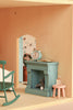 A cozy dollhouse room is brought to life with the Maileg Fully Furnished Farmhouse Set + Surprise Gift (ships in one week), featuring a teal fireplace adorned with a clock and mantel rings, alongside charming Maileg accessories. A small pink table holds a bottle beside a green rocking chair, both part of the delightful Maileg mouse furniture collection. Through the doorway, whimsical animal wallpaper and a potted plant capture the inviting spirit of this beautifully furnished farmhouse dollhouse set.