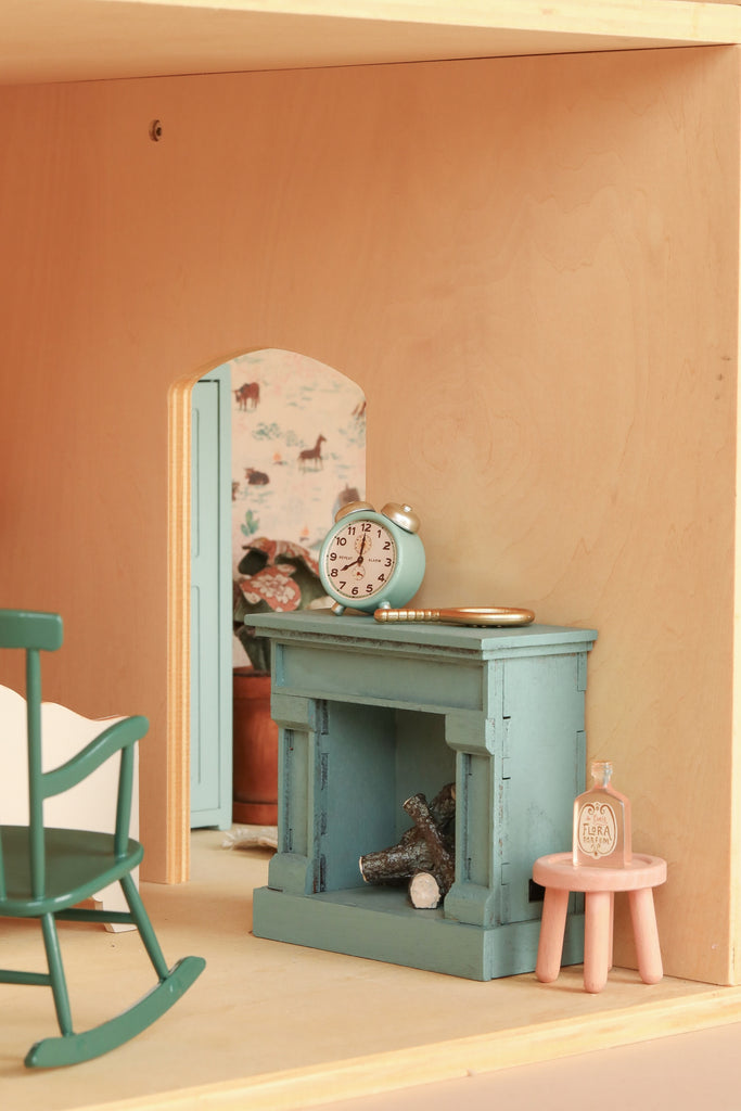A cozy dollhouse room is brought to life with the Maileg Fully Furnished Farmhouse Set + Surprise Gift (ships in one week), featuring a teal fireplace adorned with a clock and mantel rings, alongside charming Maileg accessories. A small pink table holds a bottle beside a green rocking chair, both part of the delightful Maileg mouse furniture collection. Through the doorway, whimsical animal wallpaper and a potted plant capture the inviting spirit of this beautifully furnished farmhouse dollhouse set.