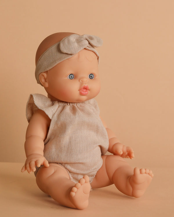 The Minikane Baby Doll (13") - Lily With Outfit features light skin, blue eyes, rosy cheeks, a beige ruffled-sleeve outfit, and a matching headband. This anatomically correct doll sits upright against a solid beige background.